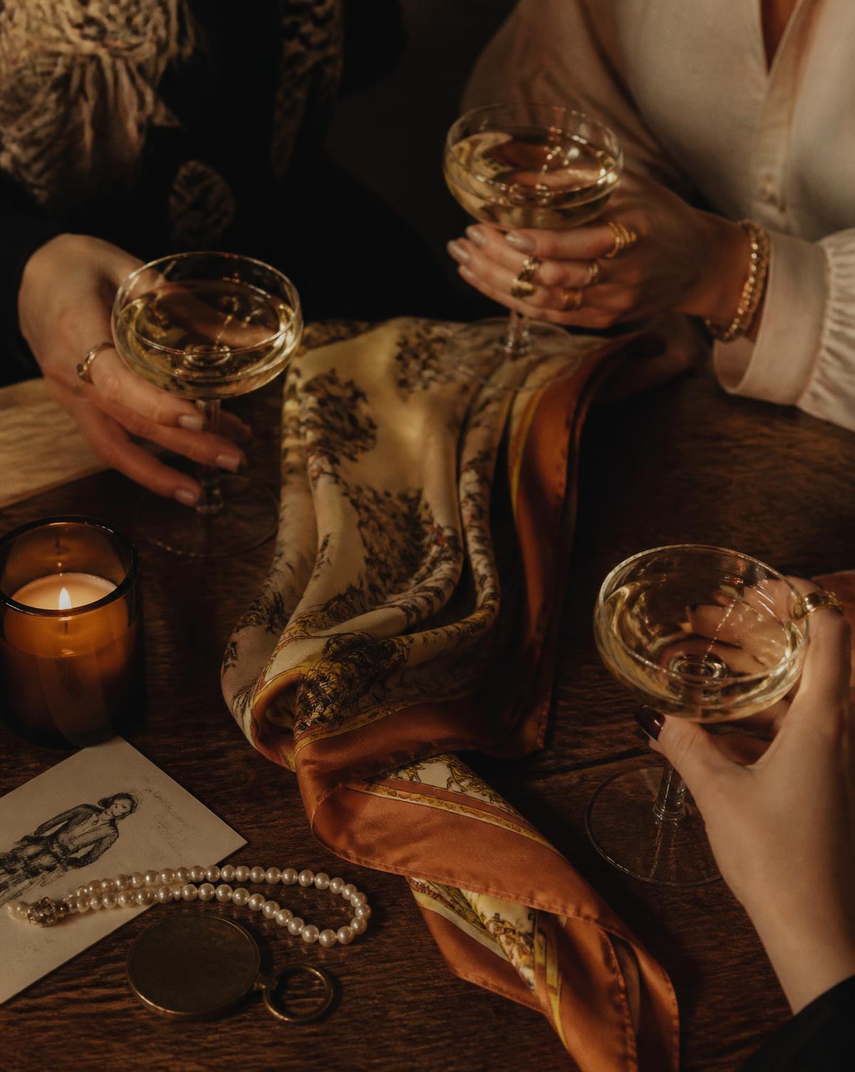 Hands holding coupe glasses over silk scarves by candlelight