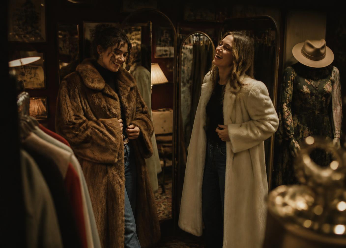 Two women trying on vintage coats in a dimly lit thrift shop after hours